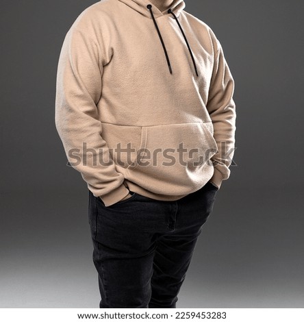 A young man in a beige hoodie and jeans stands on a gray background. Beige sweatshirt for men. Studio shooting of clothes. Men clothes. Men hoodie.