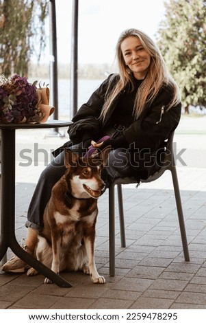 beautiful blonde girl sitting at the table on summer terrace of restaurant, funny brown dog is sitting next to her, bouquet of flowers on table, walk with pet, lake on background