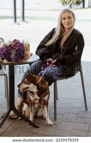 beautiful blonde girl sitting at the table on summer terrace of restaurant, funny brown dog is sitting next to her, bouquet of flowers on table, walk with pet, lake on background