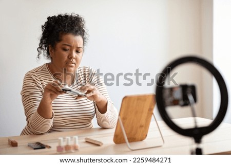 Unhappy young hispanic woman plus size beauty blogger recording video for followers, sitting at desk full of cosmetics, holding eyeshadow palette, streaming from home, using blogger set, copy space