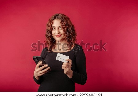 Redhead millennial woman standing isolated over red background looking at the camera with a charming, holding a phone and a credit card. Invites you to shop. An ad for a bank or mobile app.