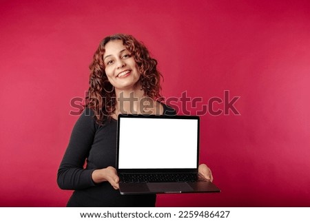 Portrait of young redhead woman isolated over red background holding with hands to a laptop with an empty white mockup screen and looks at the camera with a smile. Confident, advertising concept.