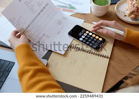 High angle of crop unrecognizable female checking financial documents and managing personal money using calculator on a smartphone while sitting at table in a cafe and having a lunch indoors. Royalty-Free Stock Photo #2259489431