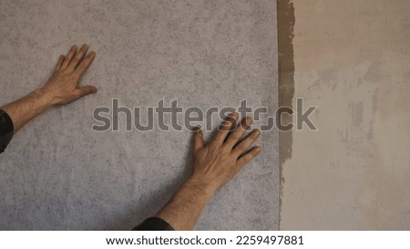 male hands touch the wall with a layer of glue applied and stick a strip of gray textured wallpaper, close-up, fragment of wallpapering in a home room with human hands Royalty-Free Stock Photo #2259497881