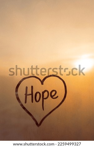 Handwritten word Hope in shape heart on misted glass on window flooded with raindrops on orange cloudy background, vertical