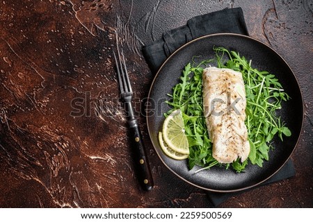 Grilled cod fish fillet served with green salad in a plate. Dark background. Top view. Copy space.