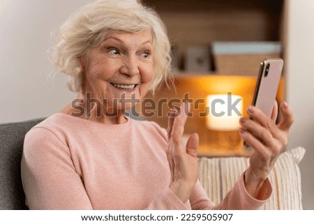 Happy mature old woman using mobile phone app for video call, waving hand, feeling excited while sitting on couch at home. Stock photo 