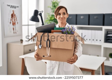 Middle age doctor woman holding cardboard box with items at the clinic smiling looking to the side and staring away thinking. 