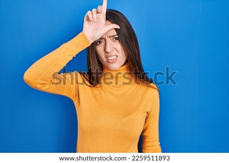 Young brunette woman standing over blue background making fun of people with fingers on forehead doing loser gesture mocking and insulting. 