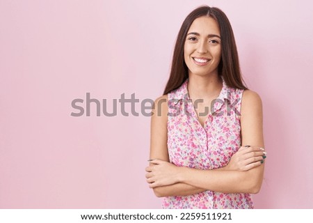 Young hispanic woman with long hair standing over pink background happy face smiling with crossed arms looking at the camera. positive person. 