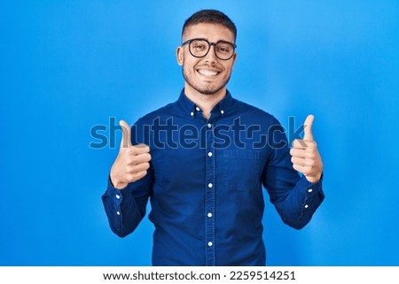 Young hispanic man wearing glasses over blue background success sign doing positive gesture with hand, thumbs up smiling and happy. cheerful expression and winner gesture.  Royalty-Free Stock Photo #2259514251
