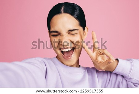 Excited young woman taking a selfie in a studio, she makes a peace sign with an expression of joy on her face. Woman having fun capturing her moments of self confidence and positivity.
