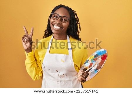 African woman holding painter palette smiling looking to the camera showing fingers doing victory sign. number two. 