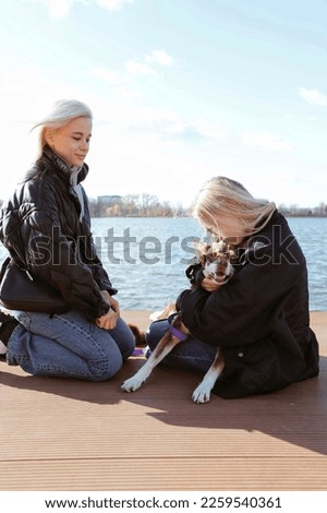 two beautiful blonde girls on pier on lake with funny brown dog, girlfriends or sisters are playing, hugging and laughing with pet, happy friends day on nature