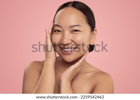 Happy Korean female with bare shoulders touching smooth face skin and looking at camera with smile against pink background