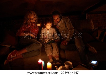 Cozy family evening. Mother, father and little girl sitting on sofa without electricity and reading book with candle light. Blackout. Hobby, leisure time. Concept of power outage, adjusting Royalty-Free Stock Photo #2259546147