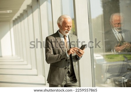 A senior business man stands in an office hallway, focused on his mobile phone. He is dressed in formal attire, exuding confidence and professionalism Royalty-Free Stock Photo #2259557865