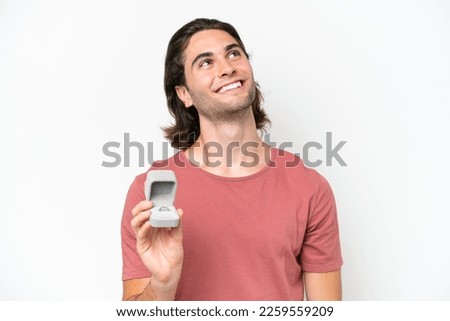 Young handsome man holding a engagement ring isolated on white background looking up while smiling