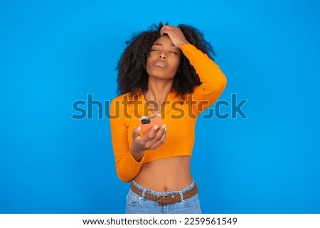 Upset depressed Young beautiful girl with wearing orange crop top over blue background makes face palm as forgot about something important holds mobile phone expresses sorrow and regret blames Royalty-Free Stock Photo #2259561549