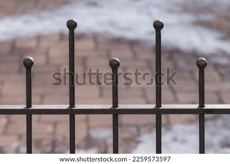 Closeup of a modern metallic fence with round-topped poles of alternating heights. Blurred brick patio with snow in the background.