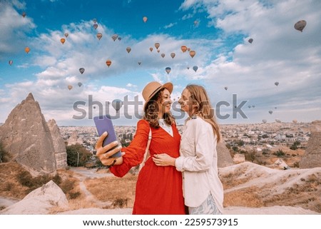 Two friends share their adventure in Cappadocia, Turkey on social networks with their smartphone selfies surrounded by breathtaking rock formations and hot air balloons.