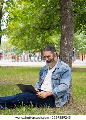 Photo of a bearded man while working online with a laptop. He sits in the park on the grass and works remotely. Vertrical photo.
