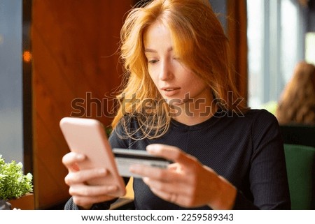 Red haired young woman making card payment through mobile phone to pay bills at a cafe. Close up.