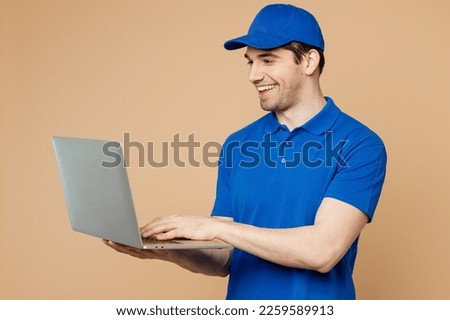 Professional delivery guy employee IT man wear blue cap t-shirt uniform workwear work as dealer courier hold use work on laptop pc computer isolated on plain light beige background. Service concept