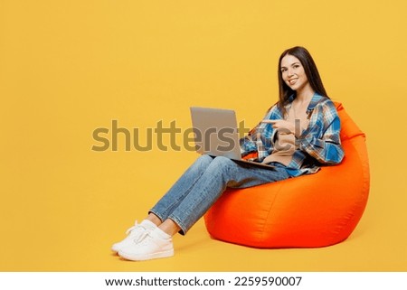 Full body young IT woman wear blue shirt beige t-shirt sit in bag chair hold use work point finger on laptop pc computer isolated on plain yellow background studio portrait. People lifestyle concept