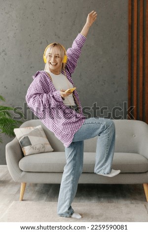 Young woman wear casual clothes headphones listen music dance raise up hand sits on grey sofa couch stay at home hotel flat rest relax spend free spare time in living room indoor People lounge concept