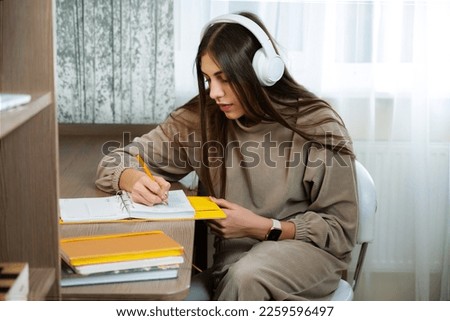 Teenage girl sits at desk at home and write in a notebook. White headphones on head for listen relax music. Thinks about plans, keeps a diary book. Royalty-Free Stock Photo #2259596497