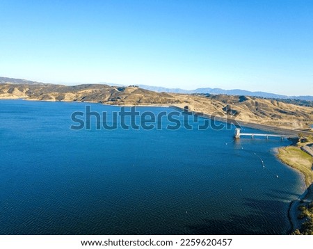 aerial shot of gorgeous summer landscape on the lake at sunset with lush green grass and trees, blue lake water and majestic mountain ranges with blue sky at Lake Castaic in California USA