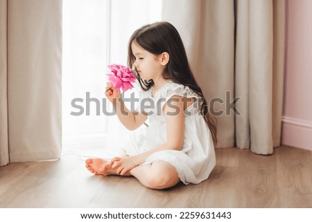 A little girl in a white dress is sitting near the window and holding a rose. A happy child at the window with a flower. Royalty-Free Stock Photo #2259631443