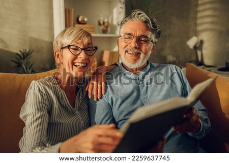 Smiling senior couple sitting on a couch at home and enjoying looking through photo album together. Happy moments, portrait of a grandparents admiring photos in family album and remembering youth.
