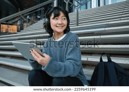 Smiling chinese student sits on stairs with tablet, draws digital art, graphic design project for freelance job, listens music in headphones and smiles.