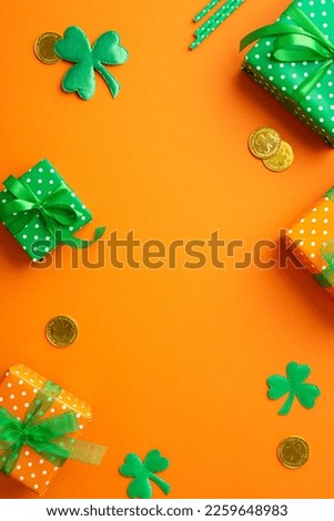 Saint Patrick's Day concept. Top view vertical photo of gift boxes with ribbon bows gold coins straws and clovers on isolated orange background with copyspace in the middle