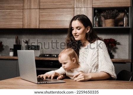 Modern childhood. Mom is sitting with toddler child and working on laptop remotely. Maternity leave for young mother. Happy family time with daughter at home