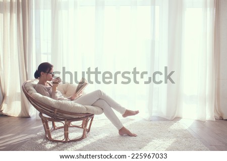 Young woman at home sitting on modern chair in front of window relaxing in her living room reading book and drinking coffee or tea Royalty-Free Stock Photo #225967033