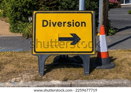 Yellow traffic diversion sign on a roadside verge with a black arrow with orange traffic cone