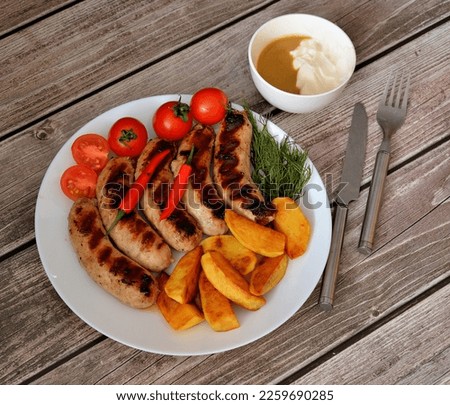 A plate with grilled sausages, cherry tomatoes, herbs, fried potatoes with hot peppers and a bowl with sauce on a wooden table. Top view, flat lay.