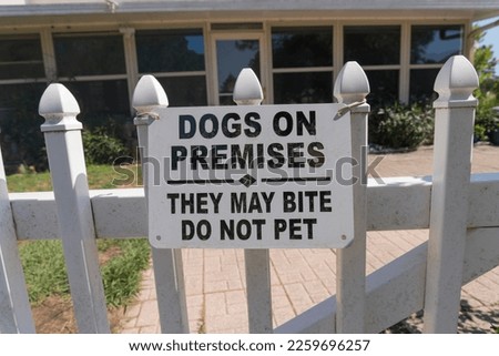 Warning sign with Dogs on Premises. They May Bite Do Not Pet hanging on a fence- Navarre, Florida. Close-up of warning sign on white picket fence against the bricks pavement and building.
