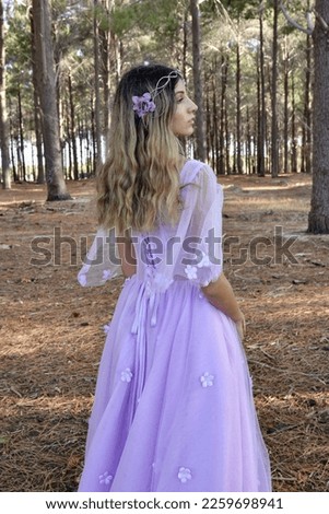 Close up portrait of beautiful young blonde model wearing a purple princess fantasy ball gown with flower crown diadem.
Pine forest location background with golden lighting.