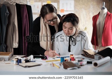 Asian middle-aged female fashion designer teaches a young teen trainee tailor in studio with colorful thread and sewing fabric for dress design collection ideas, professional boutique small business. 
