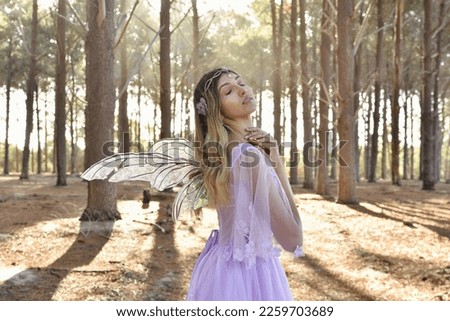 portrait of beautiful young blonde model wearing a purple princess fantasy ball gown with flower crown diadem, posing with butterfly wings. pine tree forest location background with golden lighting.