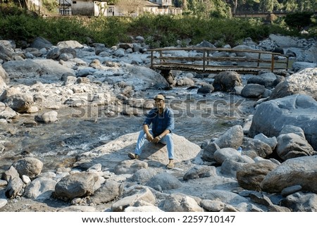 A male solo traveler sitting on a rock beside river enjoying the beauty of nature. Royalty-Free Stock Photo #2259710147