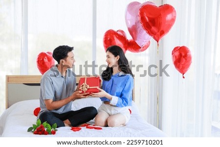 Asian young romantic cheerful lover couple male boyfriend sitting on bed giving present wrapped gift box surprise female girlfriend in bedroom decorate with pink heart shape balloon on valentine