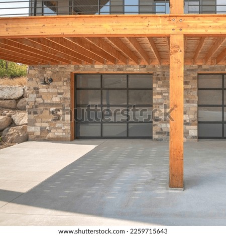 Square Two glass garage door of a modern custom house with stone veneer siding. Garage driveway under the wooden deck with metal railing against the gray siding and picture windows