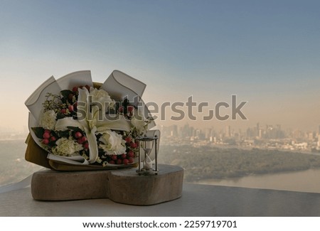 Hourglass or Sandglass is next to Bouquet of flowers placed on the timber outside the balcony with the city view background. Symbol of Romantic forever love, Free space for text.