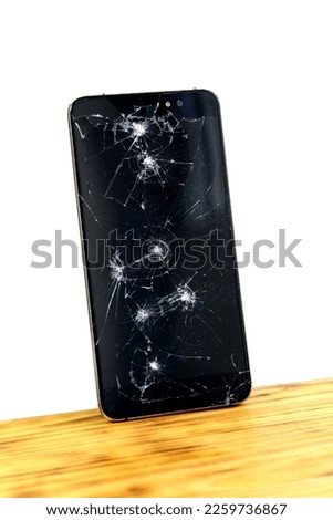 A broken cell phone stands on a wooden table tilted against a white isolated wall.