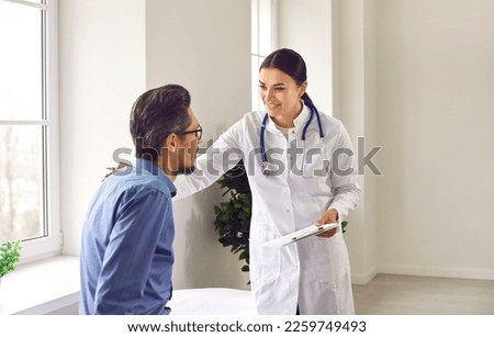 Professional female doctor approvingly, carefully puts her hand on the shoulder of young male patient. Handsome guy with glasses at doctor's appointment in modern office of medical institution. Royalty-Free Stock Photo #2259749493